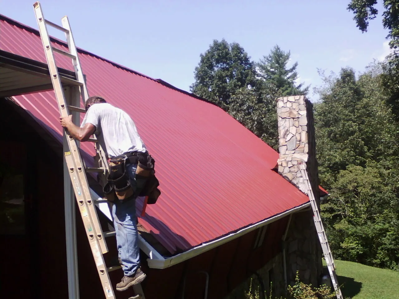 Roofer on a ladder beside a red metal roof and stone chimney — Soffit & Fascia Repair by Roofing Brothers in Troy